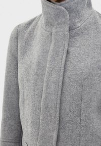 Grå ullkappa med hög krage och en strukturerad design. Har rena sömmar och en mjuk, texturerad yta. Ingen synlig hårdvara eller detaljer.