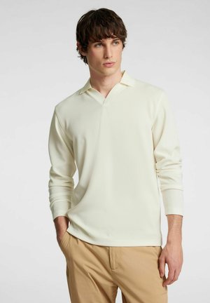 Polo a maniche lunghe color crema con scollatura a V e tessuto testurizzato, abbinato a pantaloni beige; presenta una vestibilità rilassata e dettagli di design delicati.