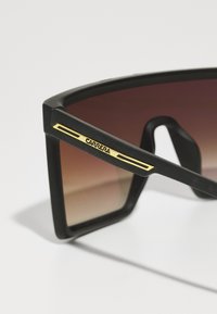 Occhiali da sole neri oversize con lenti marroni sfumate. Presentano un audace design del telaio geometrico e dettagli in oro sulle astine.