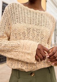 Pull en maille crochet beige avec des motifs complexes porté sur un pantalon taille haute vert olive à fermeture par boutons, mains croisées.