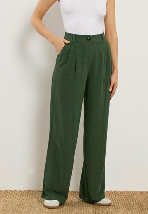 Frau trägt dunkelgrüne, hoch taillierte Weitbein-Hose mit vorderen Falten und weißen Sneakers, steht vor einer schlichten, elfenbeinfarbenen Wand.