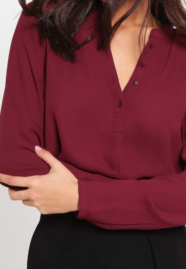 Blouse bordeaux à manches longues avec un col henley, dotée de boutons, d'une texture lisse et de manches roulées, associée à un bas noir.