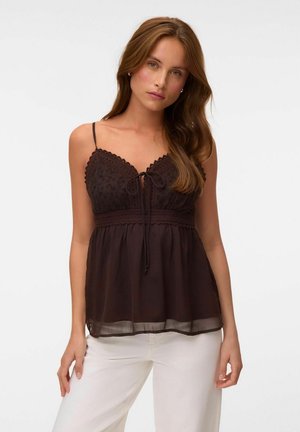 Femme aux longs cheveux bruns portant un haut en dentelle marron foncé sans manches et un pantalon blanc, debout devant un fond clair uni.