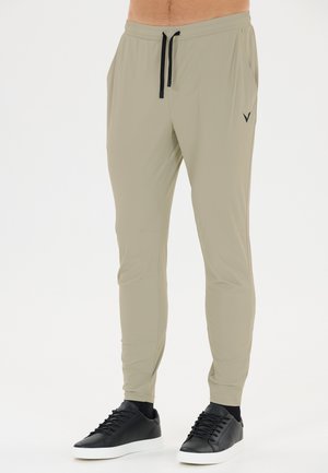 Beige tilspidsede sweatpants med sort snor båret med sorte lave sneakers med hvide såler på en person, der står på en hvid baggrund.