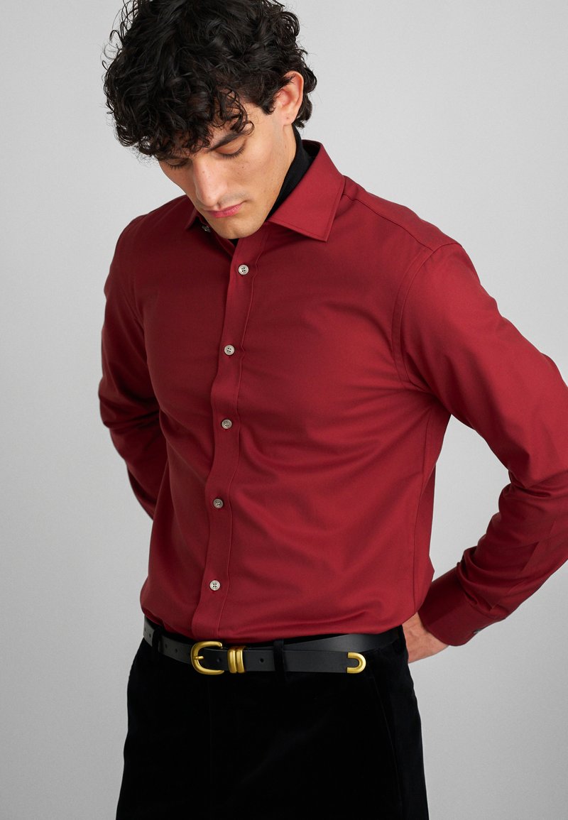 Crvena košulja s dugim rukavima i ovratnikom, s srebrnim gumbima, uparena s crnim remenom sa zlatnim detaljima.