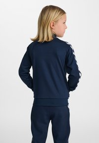 Giacca sportiva blu navy con maniche lunghe, polsini a coste, colletto alto e strisce a zigzag bianche in contrasto lungo le maniche. Tessuto morbido.