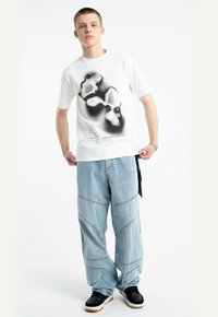 Biała bawełniana koszulka z czarnym nadrukiem graficznym, zestawiona z jasnoniebieskimi spodniami cargo z denimu, które mają widoczne szwy i luźny krój. Czarne sneakersy.