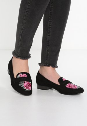 Schwarze Wildleder-Mokassins mit pinkem Rosenstick, ausgestattet mit einem dekorativen Riemen und einem niedrigen Blockabsatz. Texturiertes Material und florale Details betonen das Design.