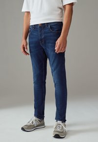 Temno modri jeans z ozkim krojem, zasnovo s petimi žepi in kontrastnimi šivi, v kombinaciji s sivo športno obutvijo.