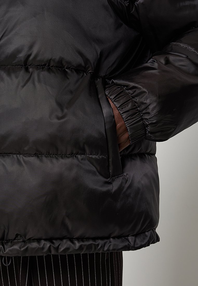 Schwarze Pufferjacke mit glänzendem Finish, elastischen Bündchen und seitlichen Taschen. Verfügt über gesteppte Abschnitte zur Isolierung und eine passgenaue Passform.