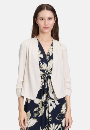 Licht beige open voorkant jasje met driekwartmouwen, gemaakt van zachte stof, gedragen over een marineblauw bloemenjurk met grote crèmekleurige bloemen.