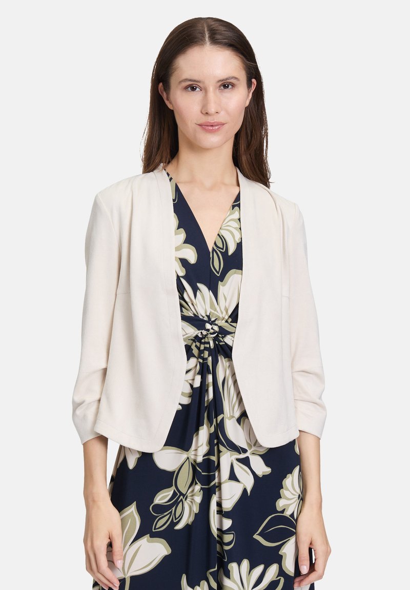 Jasno-beżowa kurtka zapinana na wierzchu z rękawami do łokci, wykonana z miękkiego materiału, noszona na granatową sukienkę w kwiaty z dużymi kremowymi kwiatami.