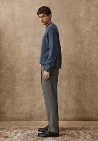 Navy blue knitted sweater, gray tailored trousers, and black loafers with hardware accents against a textured beige background.