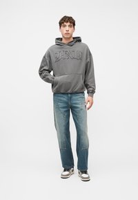 Νέος άνδρας που στέκεται, φορά γκρι φούτερ με κουκούλα και το κείμενο "FAD", μπλε τζιν με εμπρόσθιες τσέπες και άσπρα αθλητικά παπούτσια.