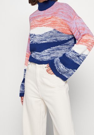 Donna che indossa un maglione a collo alto colorato con motivi blu, rosa e bianchi, abbinato a pantaloni bianchi a vita alta, mano in tasca.