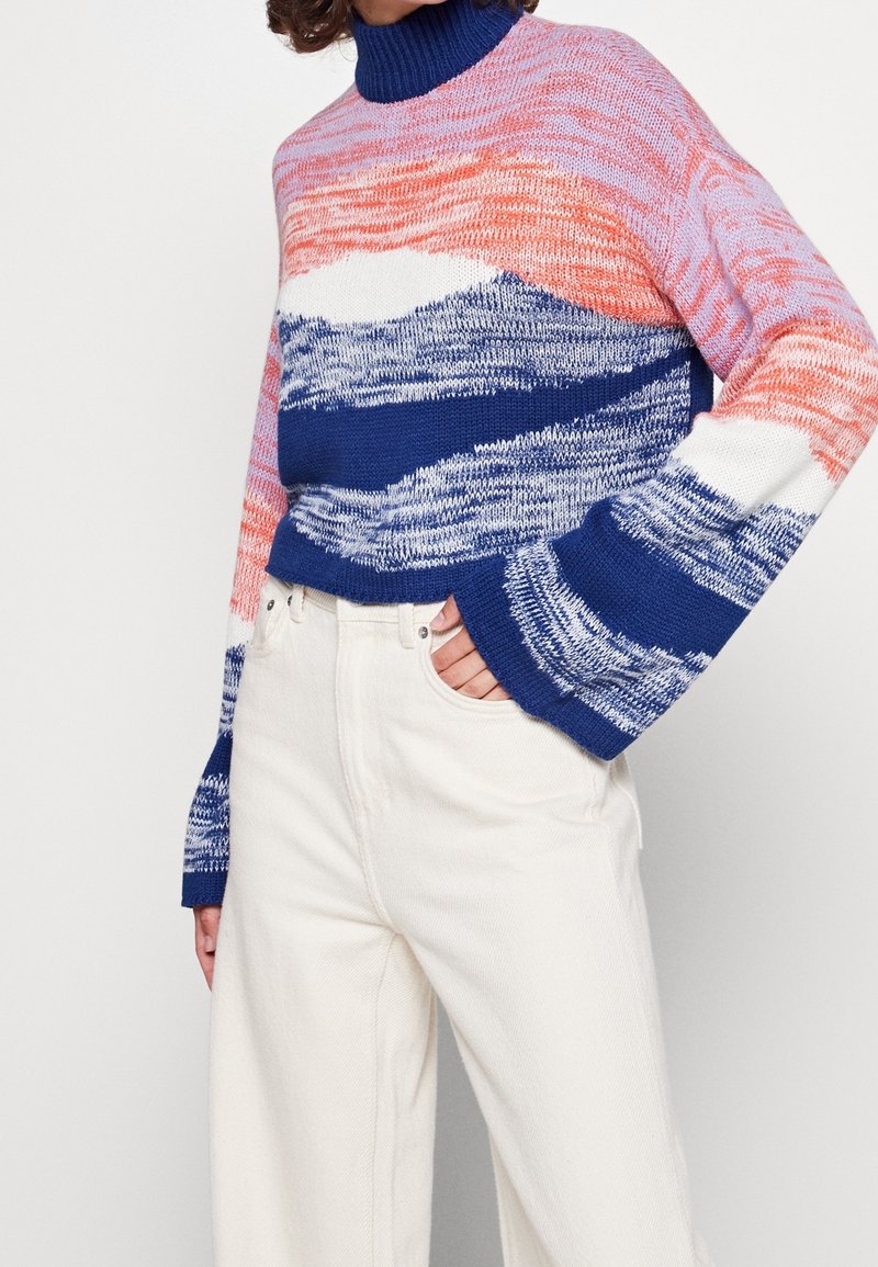 Femme portant un pull à col roulé en maille coloré avec des motifs bleus, roses et blancs, associé à un pantalon blanc taille haute, main dans la poche.