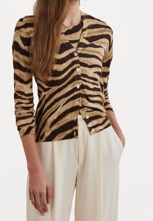 Femme portant un cardigan boutonné rayé marron et noir avec un pantalon large taille haute crème, main dans la poche.