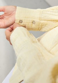 Yellow textured shirt sleeve with two metallic buttons. The fabric shows a subtle pattern, with a soft, lightweight appearance.