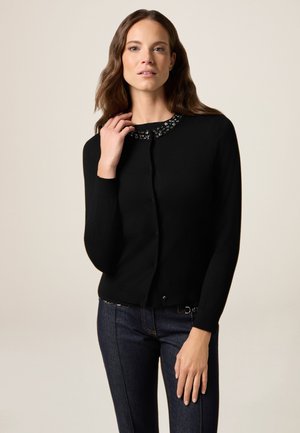 Cardigan noir avec un col rond en perles ; comporte une fermeture à boutons à l'avant et des manches longues ; associé à un jean en denim foncé. Texture lisse, design ajusté.