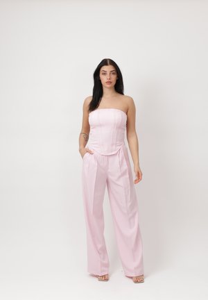 Donna con capelli lunghi e scuri che indossa un top corsetto rosa pallido senza spalline e pantaloni larghi coordinati, in piedi su uno sfondo bianco.