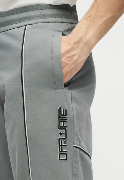 Χέρι στην τσέπη γκρι παντελονιού με ελαστική μέση και ασπρόμαυρο λογότυπο "Off-White" στο μηρό, πάνω σε απλό φόντο.