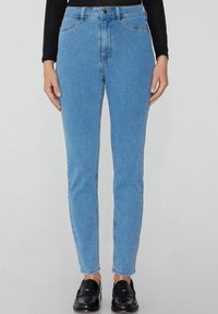 Svetle modré džíny s vysokým pasem, rovnou nohavicí, designem s pěti kapsami a viditelným kontrastním stehem. Nosené s černými mokasíny.