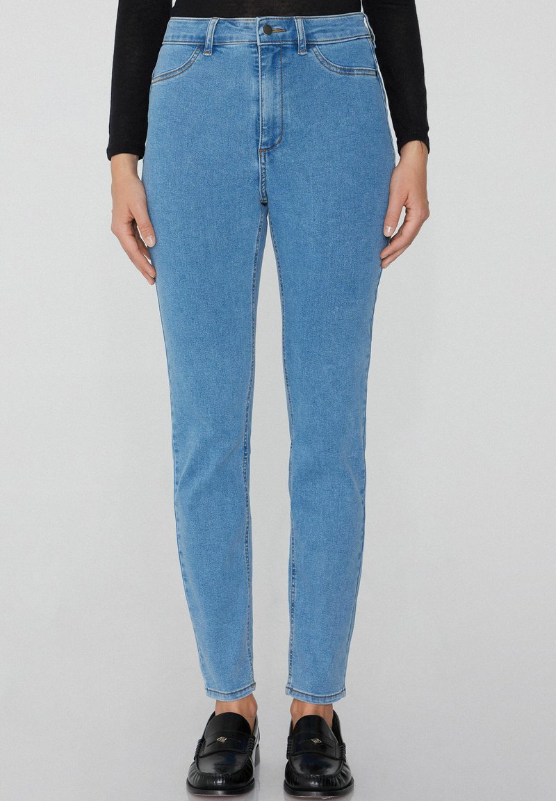 Svetle modré džíny s vysokým pasem, rovnou nohavicí, designem s pěti kapsami a viditelným kontrastním stehem. Nosené s černými mokasíny.