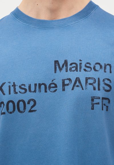 Μπλε μπλουζάκι με ξεθωριασμένο μαύρο κείμενο που γράφει «Maison Kitsuné PARIS FR 2002» στο στήθος.