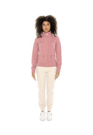 Une femme porte une veste rose à fermeture éclair, un pantalon de jogging couleur crème et des baskets blanches sur un fond blanc uni.