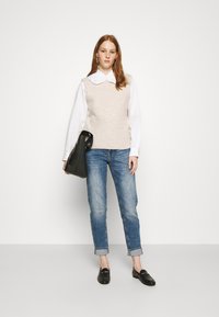 Colete de malha bege sobre uma camisa branca de colarinho, combinado com jeans claros arregaçados, mocassins pretos e uma bolsa preta, contra um fundo simples.