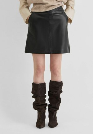Personne portant une mini-jupe en cuir noire, un pull beige et des bottes souples en daim marron foncé montant jusqu'aux genoux, debout les mains dans les poches.