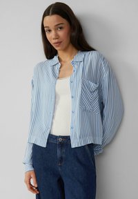 Lichtblauwe en witte gestreepte blouse met een zak, gemaakt van zachte stof. Gecombineerd met een witte tanktop en donkere spijkerbroek.