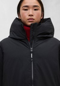 Černá péřová bunda s vysokým, strukturovaným límcem, vybavená předním zipem a značkou "ECOALF". Natažena přes červený rolák.