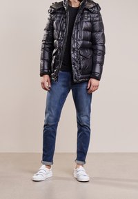 Sort pufferjakke med hætte, lynlås foran og flere lommer. Båret over en sort skjorte, parret med blå jeans og hvide sneakers.