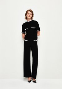 Černý zkrácený top s bílým lemováním a dvěma předními kapsami, který je kombinován s černými širokými kalhotami s vysokým pasem. Modelka má na sobě černé podpatky.