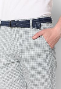 Homme portant un pantalon blanc et gris à carreaux avec une ceinture tressée bleu marine, main reposant dans la poche avant.