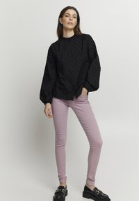 Sort tekstureret bluse med høj krave og knaplukning, parret med tætsiddende pastelfarvede lyserøde jeans og kraftige sorte loafers.