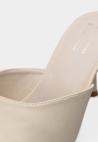 Κοντινό πλάνο ενός μπεζ δερμάτινου παπουτσιού τύπου μιουλ με τετράγωνη μύτη και τακούνι μπλοκ, που δείχνει ομαλή υφή και λεπτομέρειες ραφής.