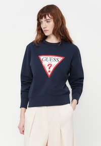 Frau mit roten Haaren, die einen dunkelblauen Sweatshirt mit einem großen roten und weißen Guess-Logo und beigen Plisseehosen trägt, die Hände in den Taschen.