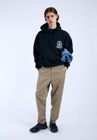 Zwarte hoodie met een witte hondenafbeelding, tan tapered broeken en zwarte sneakers. Model houdt een opgevouwen denimjack over één arm.