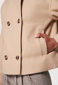 Gros plan sur une personne avec la main dans la poche, portant un manteau en laine beige avec des boutons foncés et un pantalon gris avec un détail à volants.