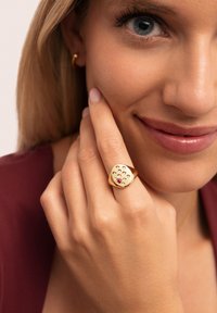 Anillo de oro con forma de cuadrado redondeado, que presenta múltiples piedras transparentes y una gema roja central. Superficie lisa y brillante, llevado en una mano.
