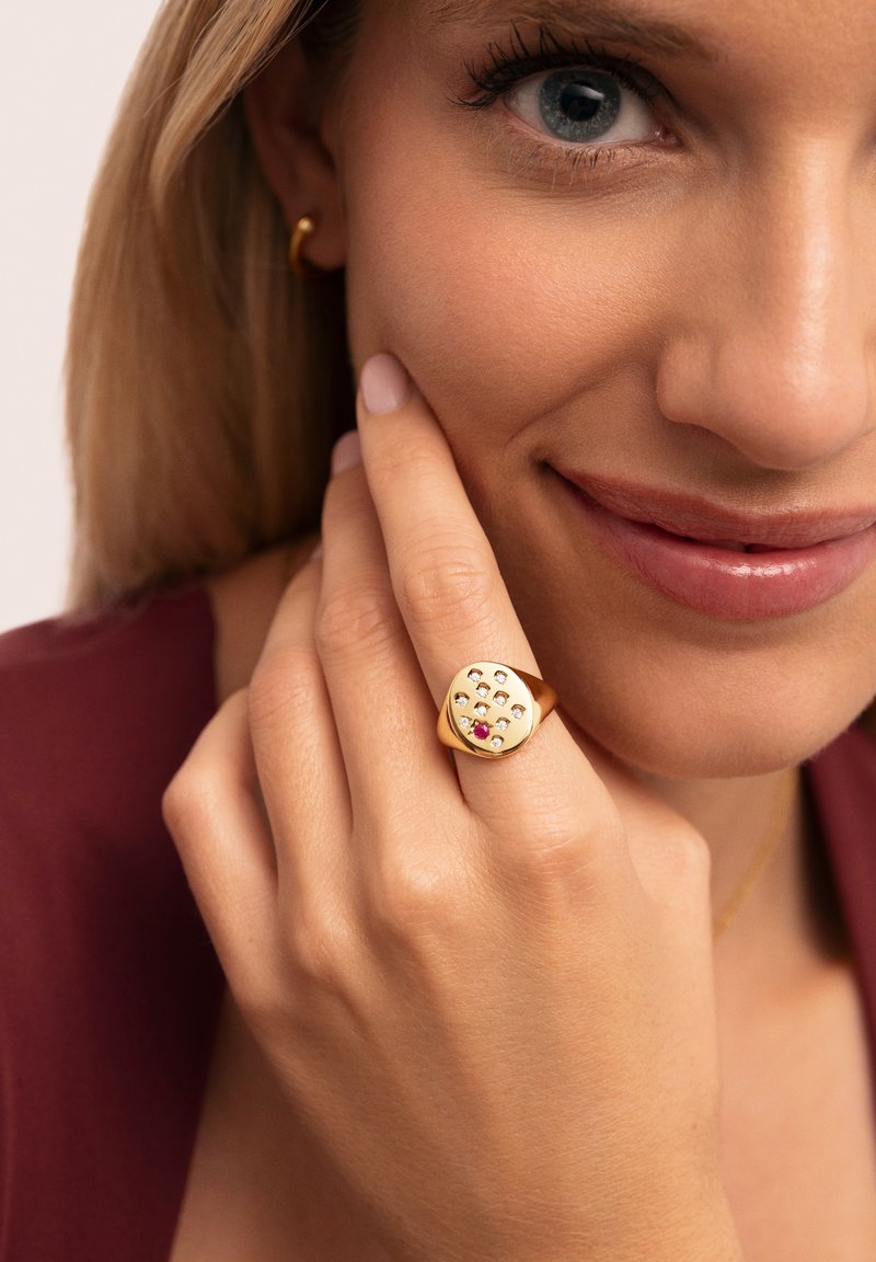 Anillo de oro con forma de cuadrado redondeado, que presenta múltiples piedras transparentes y una gema roja central. Superficie lisa y brillante, llevado en una mano.