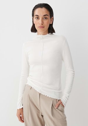 White ribbed long-sleeve top with high neck and scalloped edges, paired with light beige pinstriped trousers. Smooth texture, fitted design.