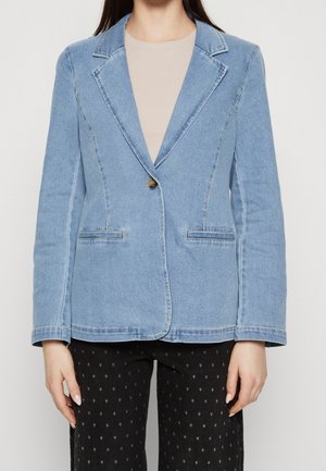 Kvinde iført en lyseblå denimblazer med en enkelt knap, beige top og sorte bukser med små stjernemønstre, stående mod en hvid baggrund.