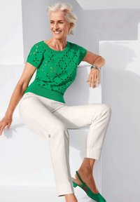Grüner Strickoberteil mit Ausschnittmuster, kombiniert mit cremefarbenen schmalen Hosen und grünen Slingback-Schuhen. Modell sitzt vor einem hellen Hintergrund.