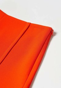 Lys orange stof med en synlig søm og en lille lynlås træk på højre kant, liggende fladt på en hvid overflade.