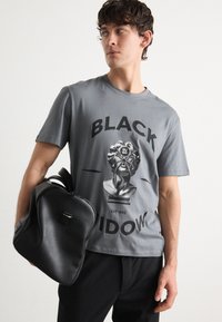 Grå bomulls t-shirt med en svart grafisk tryck av en byst med ett spindeldesign. Modellen håller en svart texturerad ryggsäck.