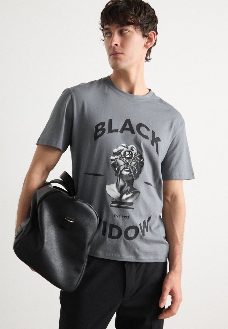 Grå bomulls t-shirt med en svart grafisk tryck av en byst med ett spindeldesign. Modellen håller en svart texturerad ryggsäck.
