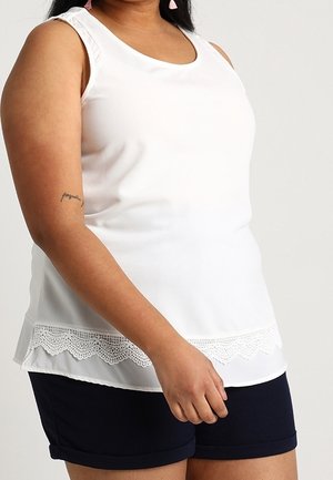Frau trägt ein weißes ärmelloses Top mit Spitzenabschluss und marineblaue Shorts, steht vor einem einfarbigen hellen Hintergrund.